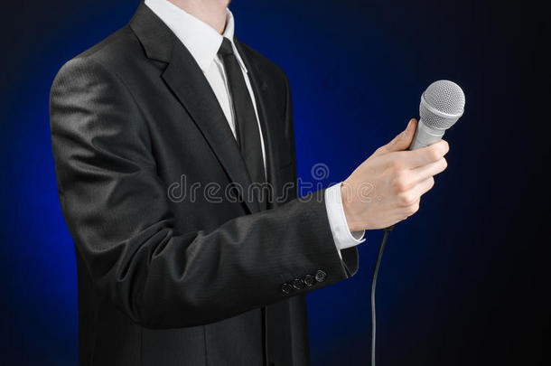 商业与演讲主题：演播室里，一个身穿黑色西装的男人在灰深蓝色的隔离背景上拿着麦克风