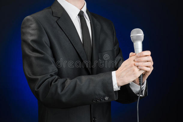 商业与演讲主题：演播室里，一个身穿黑色西装的男人在灰深蓝色的隔离背景上拿着麦克风