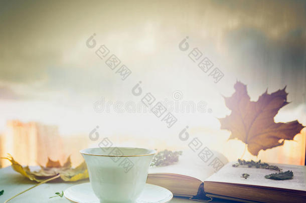 一杯茶在茶托上，茶托上有百里香，秋天的叶子和打开的书在木窗台上，在大自然的蓝天背景上