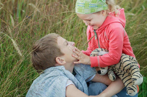 可爱的笑着蹒跚学步的女孩触摸她的兄弟兄弟的脸在夏季草地自然背景