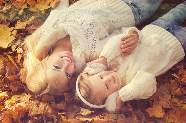 可爱微笑的母女穿着白色针织毛衣，躺在黄色的秋天树叶上休息。