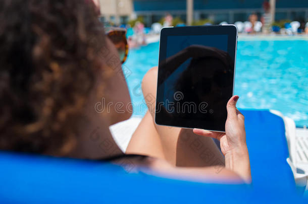 裁剪镜头的妇女阅<strong>读书籍</strong>在触摸垫，同时放松在游泳池附近的暑假