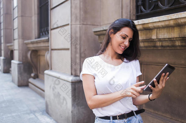 一位微笑的拉丁女人在街上行走时，在她的数字平板电脑上看照片的半长肖像