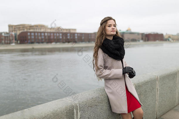 秋天站在水边的女孩。 穿着黑色外套、围巾和红色连衣裙的女孩映衬着灰色的天空。 不是孤立的。