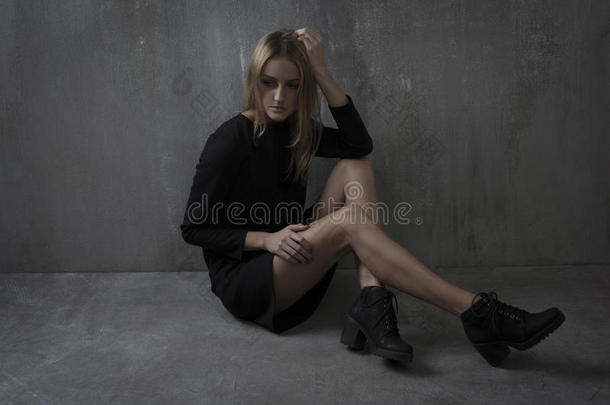 时尚的女人穿着黑色的连衣裙，坐在灰色的背景上，质感的墙壁上