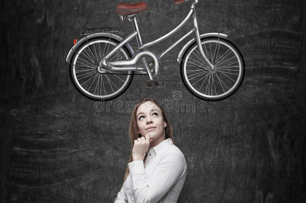 一个穿着正式衣服的女孩正在梦想一辆新自行车。 自行车的草图画在黑板上。