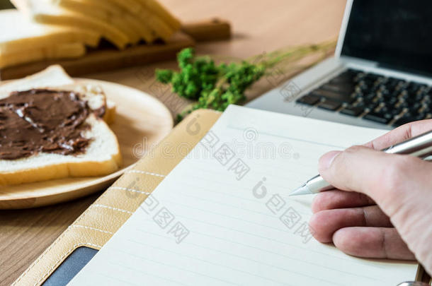 一个人拿着笔在笔记本上。 工作台包括笔记本电脑、笔记本电脑和带巧克力切片面包的早餐