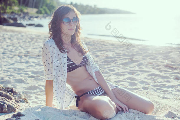 海滩女人戴太阳镜暑假旅行假期。 穿着衬衫和比基尼的女孩坐在沙滩上