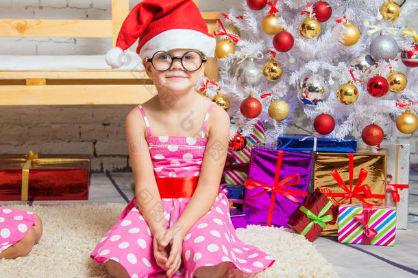 戴着红色帽子和有趣的圆形眼镜的女孩坐在圣诞树的垫子上
