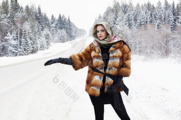 穿着毛皮大衣的漂亮女孩在森林里的一条冬天的路上等着车