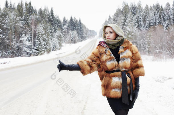 穿着毛皮大衣的漂亮女孩在森林里的一条冬天的路上等着车