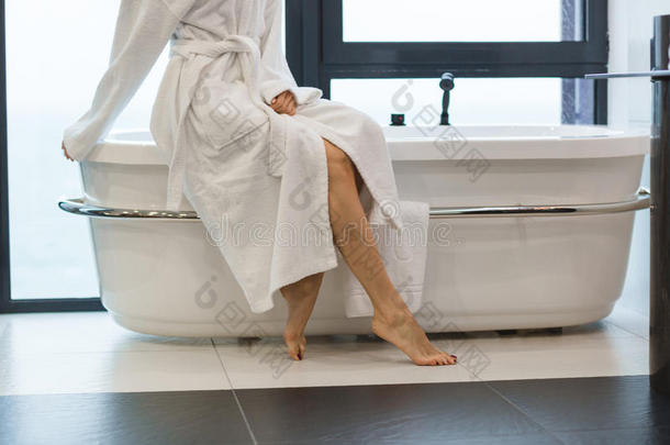 迷人的年轻光脚女人穿着白色浴袍坐在浴缸上