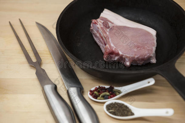 一片美味的新鲜生猪肉特写叉子，刀，香料，洋葱，大蒜，在桌上乡村厨房的铸铁煎锅上