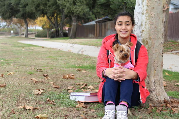 少数民族十几岁的女孩在公园的一棵树下玩狗，带着书和学校的背包