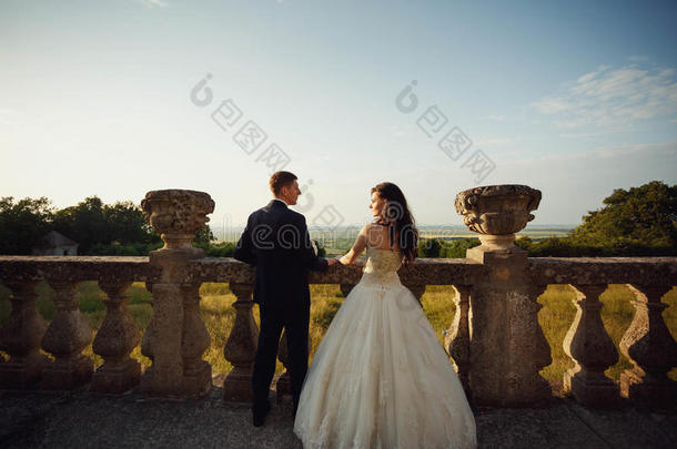 漂亮的新婚夫妇，时尚的新娘和英俊的新郎，拥抱在城堡附近，背景有一块田野