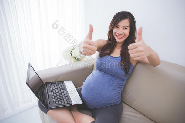 美丽的亚洲孕妇坐在沙发上，<strong>上面</strong>有<strong>笔记本</strong>电脑