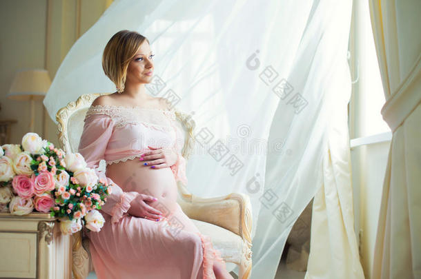 可爱的孕妇坐在椅子上，靠近<strong><strong>滚滚</strong></strong>的窗帘，用爱拥抱腹部