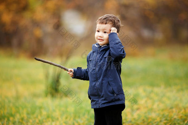可爱的男孩手里拿着木棍在秋天的公园户外玩耍。 困惑，想着该怎么办，看着一边