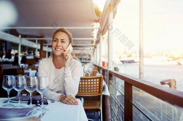 迷人的微笑的女人在温暖的秋日坐在现代的人行道咖啡馆里着手机，