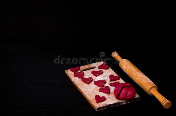 面团和生红色心形饼干在木板上与面粉，木制擀面杖。 黑色桌子