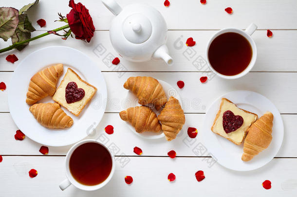 情人节为夫妇提供早餐，包括烤面包、心形果酱、牛角面包、红玫瑰花、花瓣和茶