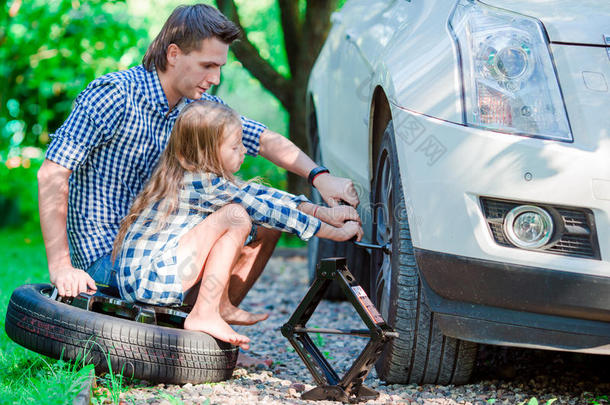 可爱的小女孩坐在轮胎上，帮助父亲在美丽的夏天在户外更换汽车车轮