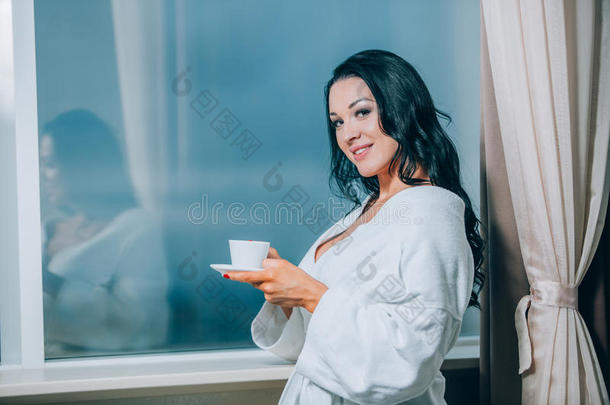 用新鲜咖啡取暖。 穿着白色浴袍的漂亮年轻女人喝着咖啡，透过窗户看着