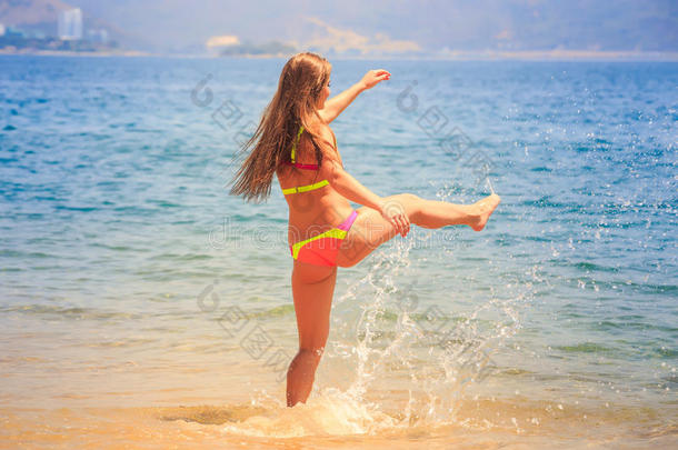 穿着比基尼的金发苗条女孩踏入海滩上的大海飞溅