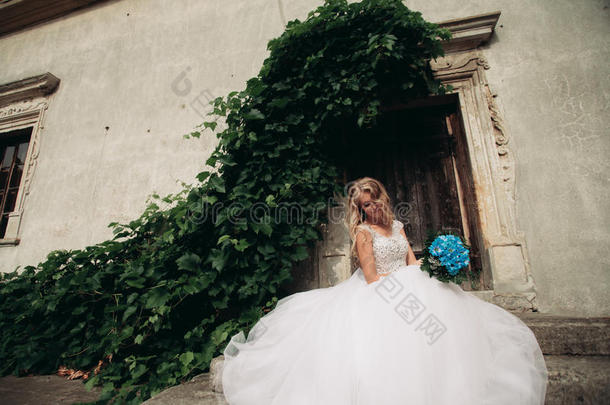 美丽的年轻金发新娘，新娘的新娘花束坐在楼梯下华丽的植物
