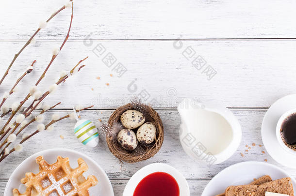 复活节早餐，包括鹌鹑蛋、华夫饼、水果果酱、牛奶、咖啡和三明治，白色木制背景上有柳树枝