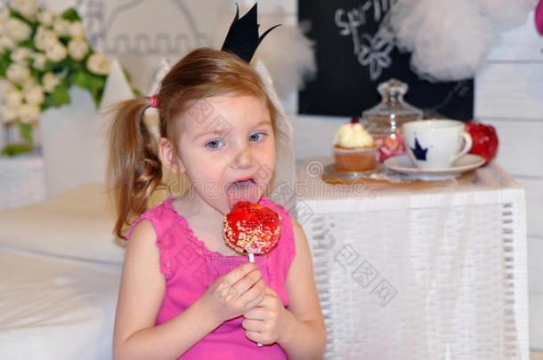 一个小女孩，头上戴着皇冠，在儿童房里舔着一根亮晶晶的焦糖