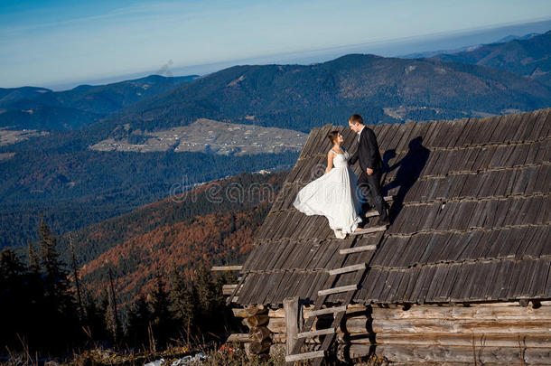 漂亮的新婚夫妇站在乡间<strong>房</strong>子的屋顶上。 惊人的山地景观背景。 <strong>蜜月</strong>