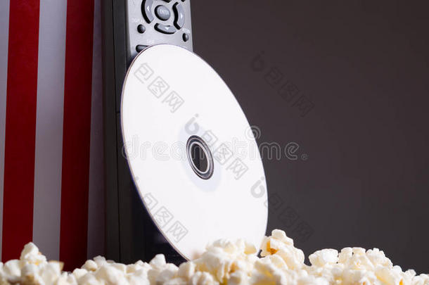 特写红色白色条纹容器站起来，爆米花躺在周围，DVD光盘和遥控器靠在盒子上