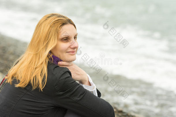 在寒冷的天气里，一个女孩坐在海滩上，在阴天里沉思地看着远方