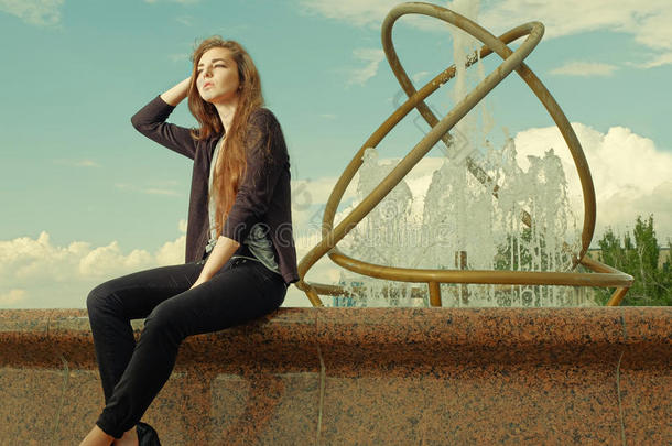 漂亮的少女坐在花岗岩喷泉上。 城市户外，青少年的街头生活方式。 色调的图像。