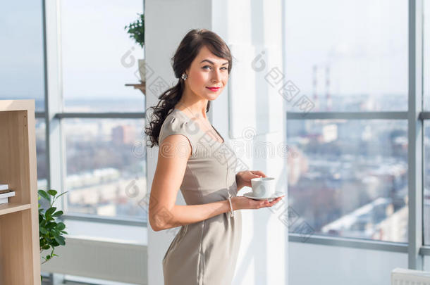 优雅的年轻女人站在一个宽敞的灯光工作室里，喝着咖啡，微笑着，在大窗户附近做梦