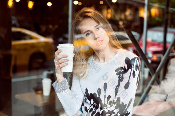 美丽的白种人女人坐在靠近窗户的城市咖啡店里，喝着纸杯里的咖啡，看着相机