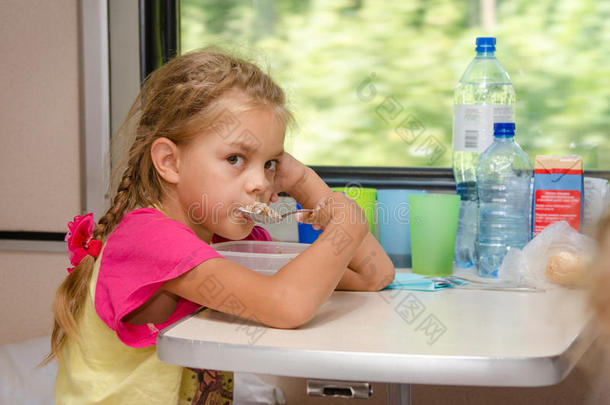 一个六岁的女孩坐在火车上，坐在汽车二等车厢的较低位置的桌子上，吃粥