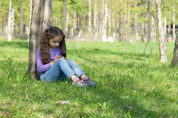 可爱的长发女孩坐在公园的草地上，手里拿着手机，用手机发送信息