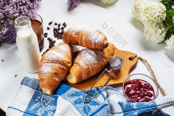 早餐有新鲜的牛角面包，一杯咖啡，牛奶，草莓酱和白色木制背景上的花