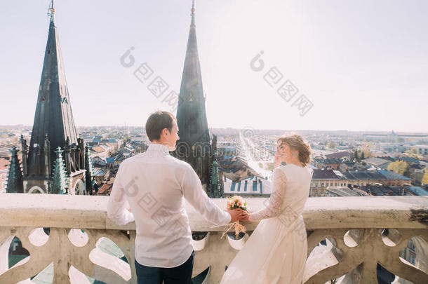 穿着白色连衣裙的美丽新娘和英俊的新郎站在阳台上眺望城市