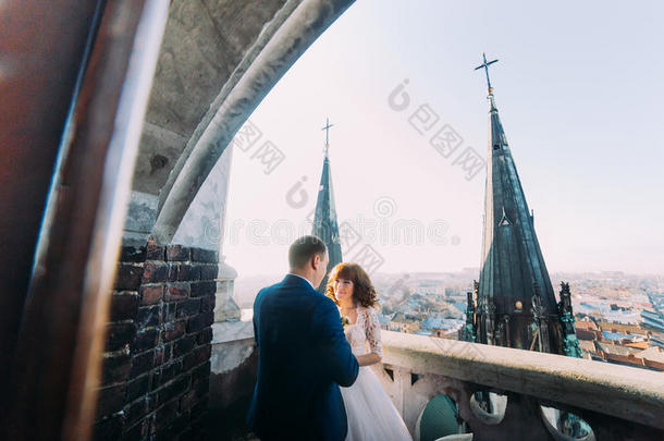 温柔美丽的新娘和新郎牵手拥抱面对面的古老阳台，背景城市景观