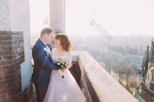 温柔美丽的新娘和新郎牵手拥抱面对面的古老阳台，背景城市景观
