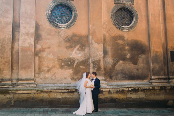 华丽的新娘穿着白色连衣裙，英俊的新郎面对面地拿着新娘的花束，靠近老教堂的棕色墙壁