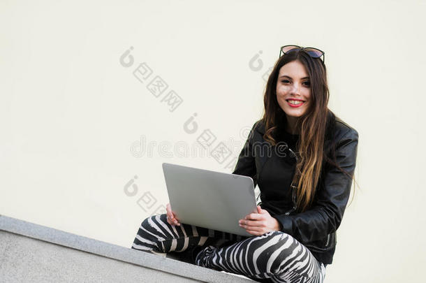 美丽的学生女人正在用笔记本电脑微笑，坐在大学校园的老墙上。 漂亮的女孩
