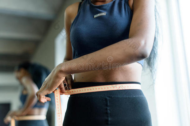 穿着运动服装的年轻成年黑人妇女的特写。 非裔美国女孩测量腰部与黄色胶带关闭
