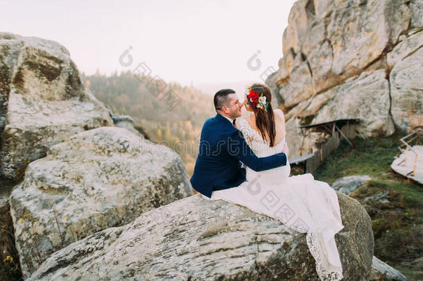 优雅的新郎穿着时尚的蓝色西装，慈爱地抱着穿着白色衣服的新娘，在雄伟的山岩上戴着可爱的头圈