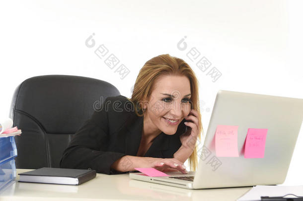 金发女商人在办公室笔记本电脑办公桌上工作，用手机微笑说话