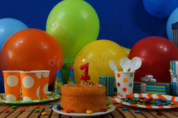 乡村木桌上的生日蛋糕，背景是五颜六色的气球、礼物、塑料杯和带糖果的塑料盘子