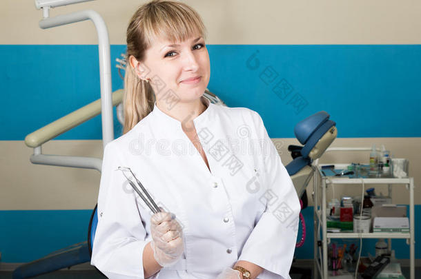 美丽的金发女子牙医拿着一件医疗器械。 背景中的牙科设备